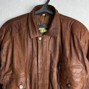 May include: A brown leather jacket with a collared neckline and two front pockets. The jacket has a button closure and a zipper pocket on the right side. The label reads "AVAL CONT".