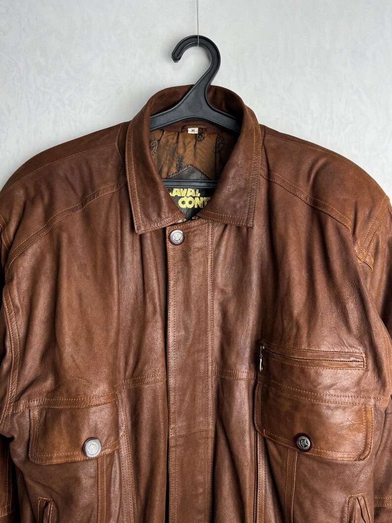 May include: A brown leather jacket with a collared neckline and two front pockets. The jacket has a button closure and a zipper pocket on the right side. The label reads "AVAL CONT".
