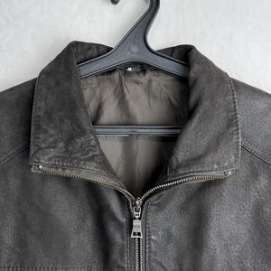 Puede incluir: Chaqueta de cuero gris oscuro con cierre de cremallera completo y cuello. La chaqueta est&aacute; colgada de una percha de pl&aacute;stico negro. El cuero tiene una apariencia texturizada.