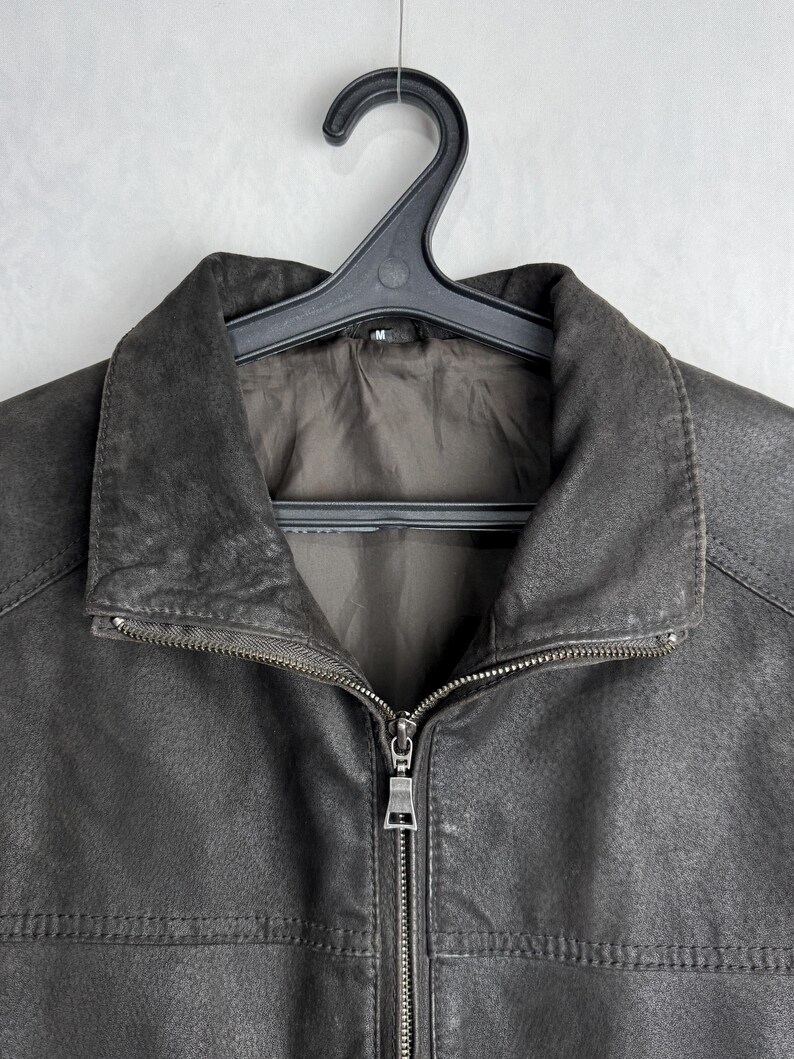 Puede incluir: Chaqueta de cuero gris oscuro con cierre de cremallera completo y cuello. La chaqueta est&aacute; colgada de una percha de pl&aacute;stico negro. El cuero tiene una apariencia texturizada.
