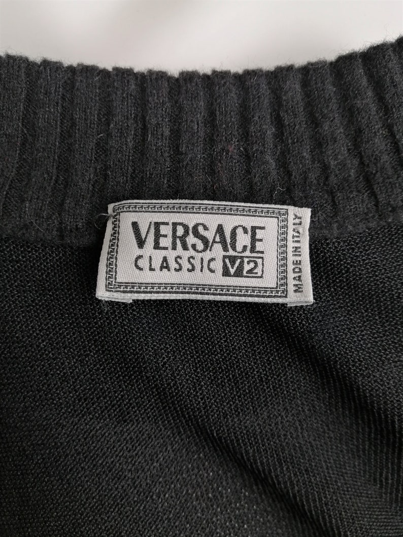 以下が含まれることがあります： 黒いニット生地に、「VERSACE CLASSIC V2 MADE IN ITALY」と書かれた白いラベルが付いている。