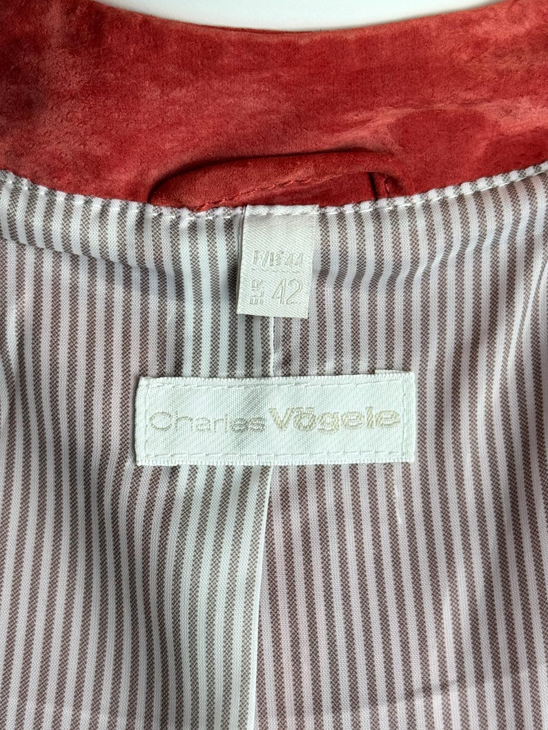 Puede incluir: Primer plano del interior de una chaqueta de ante roja. El forro es blanco con finas rayas verticales marrones. Una etiqueta blanca con el texto "Charles V&ouml;gele" est&aacute; cosida en el forro. Una etiqueta m&aacute;s peque&ntilde;a muestra la talla "42".