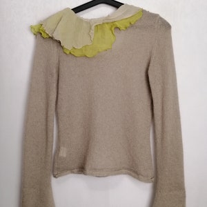 Puede incluir: Un su&eacute;ter de punto beige con mangas largas y un cuello con volantes hecho de una tela superpuesta en tonos crema y verde.