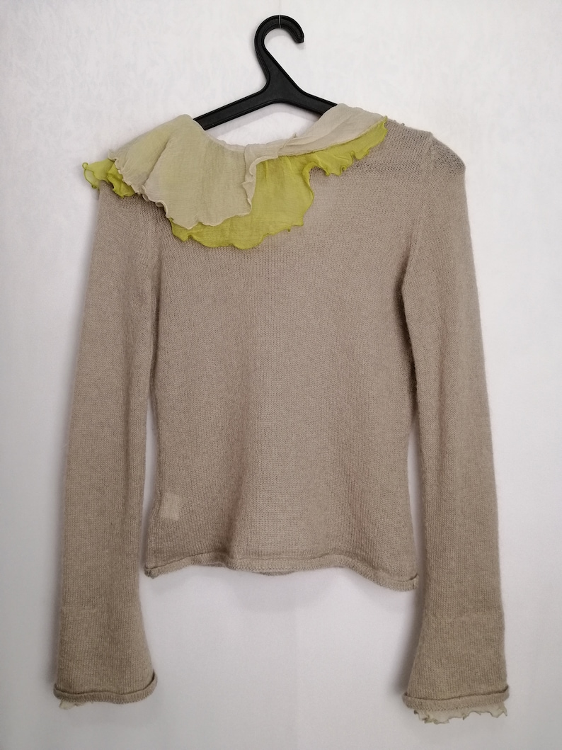 Puede incluir: Un su&eacute;ter de punto beige con mangas largas y un cuello con volantes hecho de una tela superpuesta en tonos crema y verde.