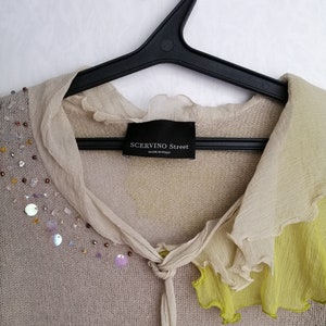 Puede incluir: Un c&aacute;rdigan de punto beige con cuello de volantes y ribete amarillo. El c&aacute;rdigan tiene cierre de nudo y est&aacute; decorado con lentejuelas y cuentas. La etiqueta dice "SCERVINO Street Made in Italy".
