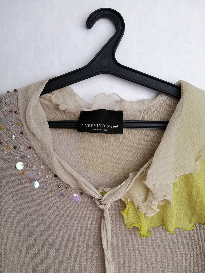 Puede incluir: Un c&aacute;rdigan de punto beige con cuello de volantes y ribete amarillo. El c&aacute;rdigan tiene cierre de nudo y est&aacute; decorado con lentejuelas y cuentas. La etiqueta dice "SCERVINO Street Made in Italy".