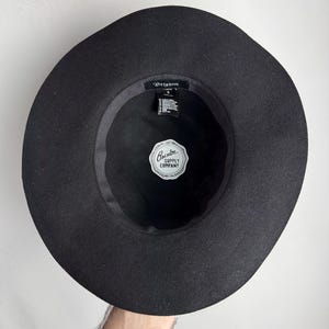 Puede incluir: Sombrero fedora de fieltro negro con ala ancha. El sombrero tiene una banda negra con una etiqueta blanca que dice "Brixton Supply Company".