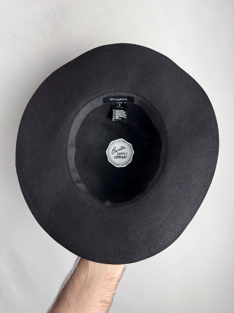 Puede incluir: Sombrero fedora de fieltro negro con ala ancha. El sombrero tiene una banda negra con una etiqueta blanca que dice "Brixton Supply Company".