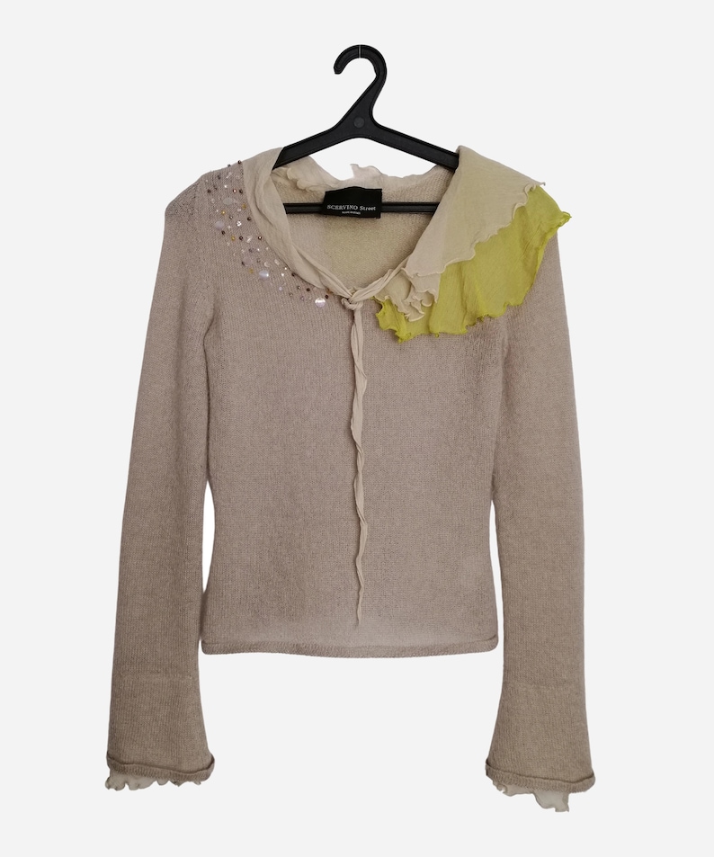 Puede incluir: Un su&eacute;ter de punto beige con un cuello de volantes en tonos blancos y amarillos. El su&eacute;ter tiene una decoraci&oacute;n de cuentas alrededor del escote y un lazo en el cuello.