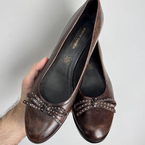 Puede incluir: Un par de bailarinas de cuero marr&oacute;n con lazos decorativos. Las bailarinas tienen una punta redondeada y un peque&ntilde;o lazo con tachuelas plateadas. La marca "Pierre Cardin" es visible en la plantilla.
