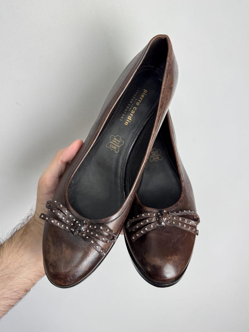 Puede incluir: Un par de bailarinas de cuero marr&oacute;n con lazos decorativos. Las bailarinas tienen una punta redondeada y un peque&ntilde;o lazo con tachuelas plateadas. La marca "Pierre Cardin" es visible en la plantilla.