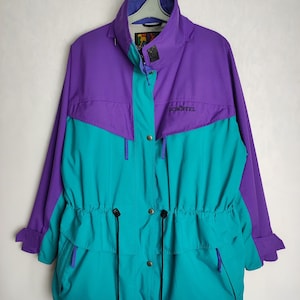 Op de afbeelding: Een paarse en turquoise blauwe jas met een trekkoord in de taille en het woord "Schöffel" geborduurd op de voorkant.