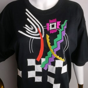 May include: Black t-shirt with a colourful abstract design embroidered on the front. The design features geometric shapes, lines, and swirls in shades of white, red, orange, yellow, green, purple, and pink.