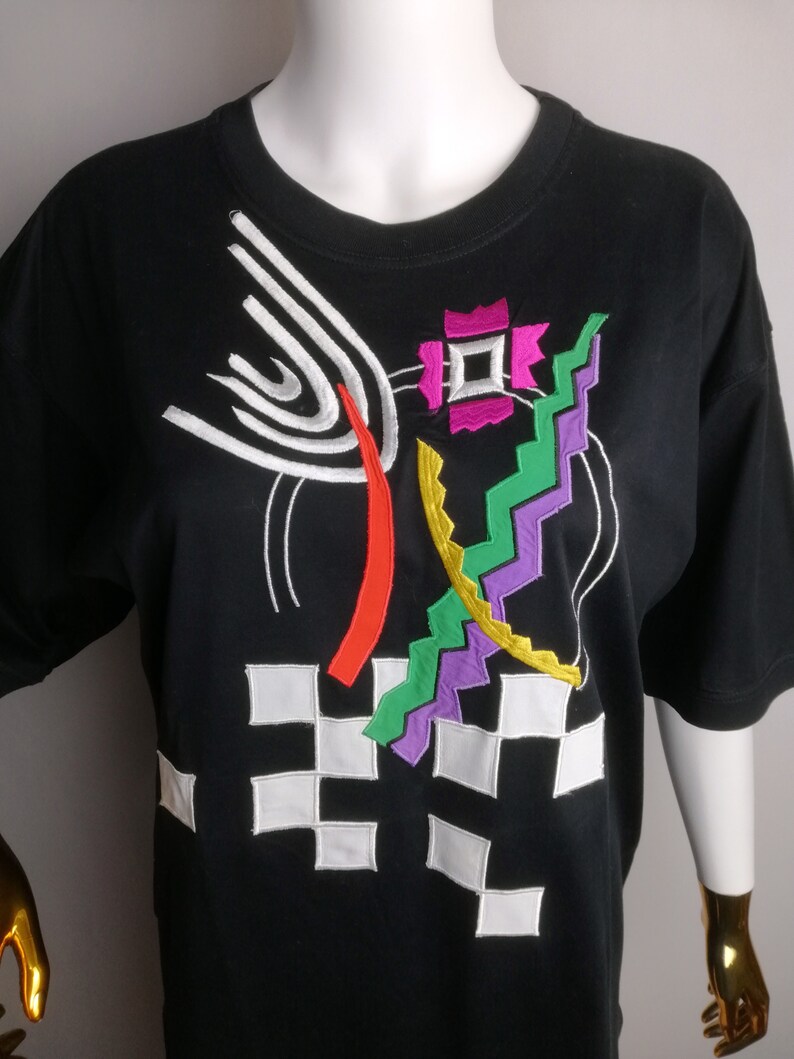 May include: Black t-shirt with a colourful abstract design embroidered on the front. The design features geometric shapes, lines, and swirls in shades of white, red, orange, yellow, green, purple, and pink.