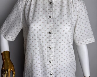 Pode incluir: Uma camisa branca de manga curta com botões e um pequeno padrão repetido de flores vermelhas e pretas. A camisa tem uma gola clássica e um fecho de botões na frente.