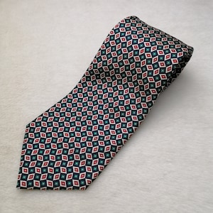 May include: A patterned necktie with a repeating geometric design. The tie features a navy blue background with a pattern of red, teal, and white diamond shapes. The tie is made of a smooth fabric and is suitable for formal occasions.