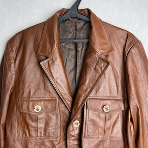 May include: A brown leather jacket with a classic design, featuring a notched collar, two chest pockets, and two lower pockets. The jacket has gold-colored buttons and is hanging on a black hanger.