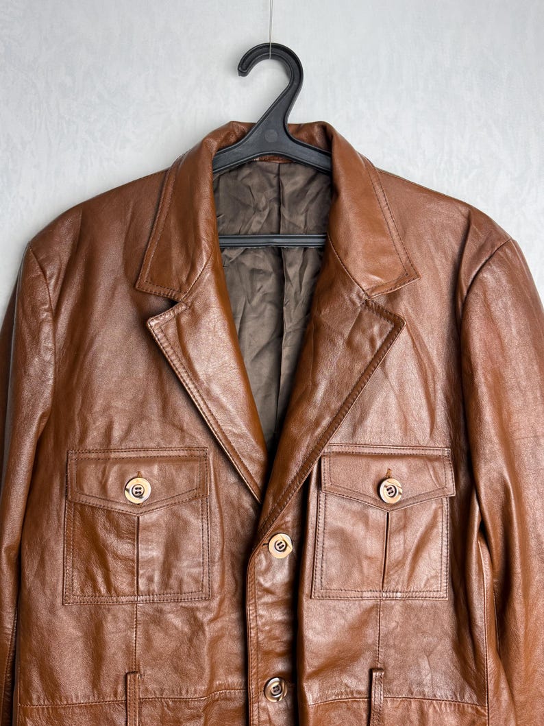 May include: A brown leather jacket with a classic design, featuring a notched collar, two chest pockets, and two lower pockets. The jacket has gold-colored buttons and is hanging on a black hanger.