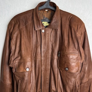 May include: A brown leather jacket with a collared neckline and multiple pockets. The jacket has a button-up front and decorative buttons on the pockets. The jacket is hanging on a black hanger.