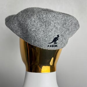 Pode incluir: Boina Kangol cinza com o logotipo do canguru preto e o nome da marca. O chap&eacute;u &eacute; feito de um tecido texturizado e &eacute; exibido em uma cabe&ccedil;a de manequim dourada. Um acess&oacute;rio cl&aacute;ssico para qualquer guarda-roupa.