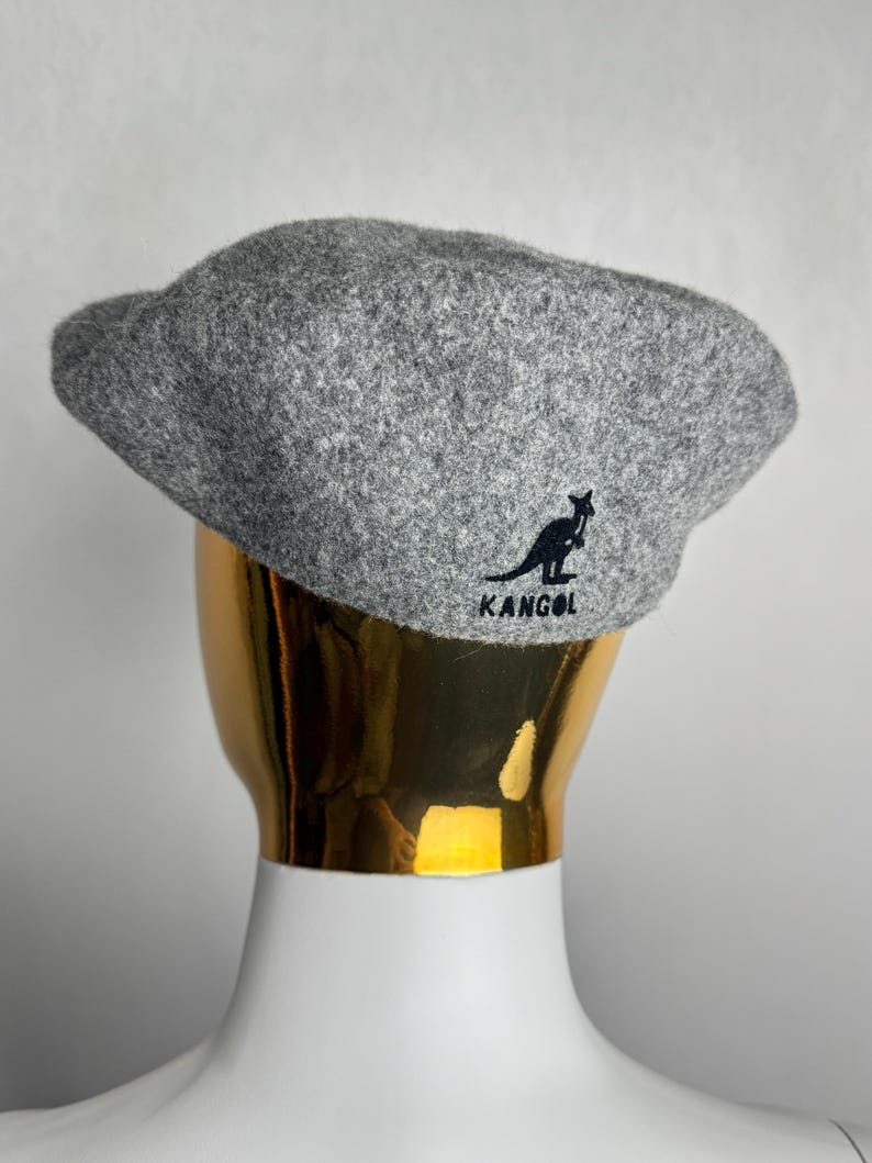 Pode incluir: Boina Kangol cinza com o logotipo do canguru preto e o nome da marca. O chap&eacute;u &eacute; feito de um tecido texturizado e &eacute; exibido em uma cabe&ccedil;a de manequim dourada. Um acess&oacute;rio cl&aacute;ssico para qualquer guarda-roupa.