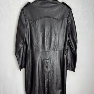 May include: A black leather coat hanging on a black hanger. The coat has a long length, button closure, and shoulder epaulets. The coat is made of a textured leather material.
