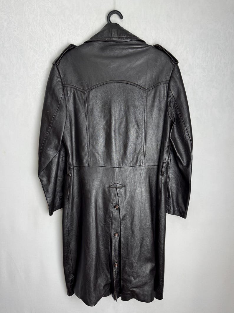 May include: A black leather coat hanging on a black hanger. The coat has a long length, button closure, and shoulder epaulets. The coat is made of a textured leather material.
