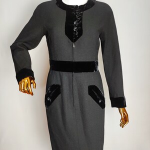 K&ouml;nnte beinhalten: Ein schwarzes Kleid mit Samtbesatz und einem Rei&szlig;verschluss vorne. Das Kleid hat eine figurbetonte Silhouette und einen G&uuml;rtel.