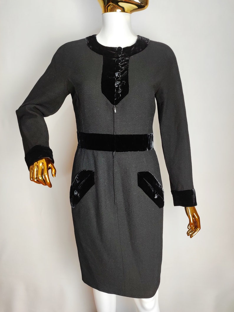K&ouml;nnte beinhalten: Ein schwarzes Kleid mit Samtbesatz und einem Rei&szlig;verschluss vorne. Das Kleid hat eine figurbetonte Silhouette und einen G&uuml;rtel.