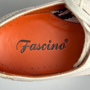 K&ouml;nnte beinhalten: Nahaufnahme eines cremefarbenen Schuhs mit wei&szlig;en Schn&uuml;rsenkeln. Der Schuh hat eine orangefarbene Innensohle mit dem Wort "Fascino" in Schreibschrift. Der Schuh scheint aus Leder zu sein und hat dekorative N&auml;hte.