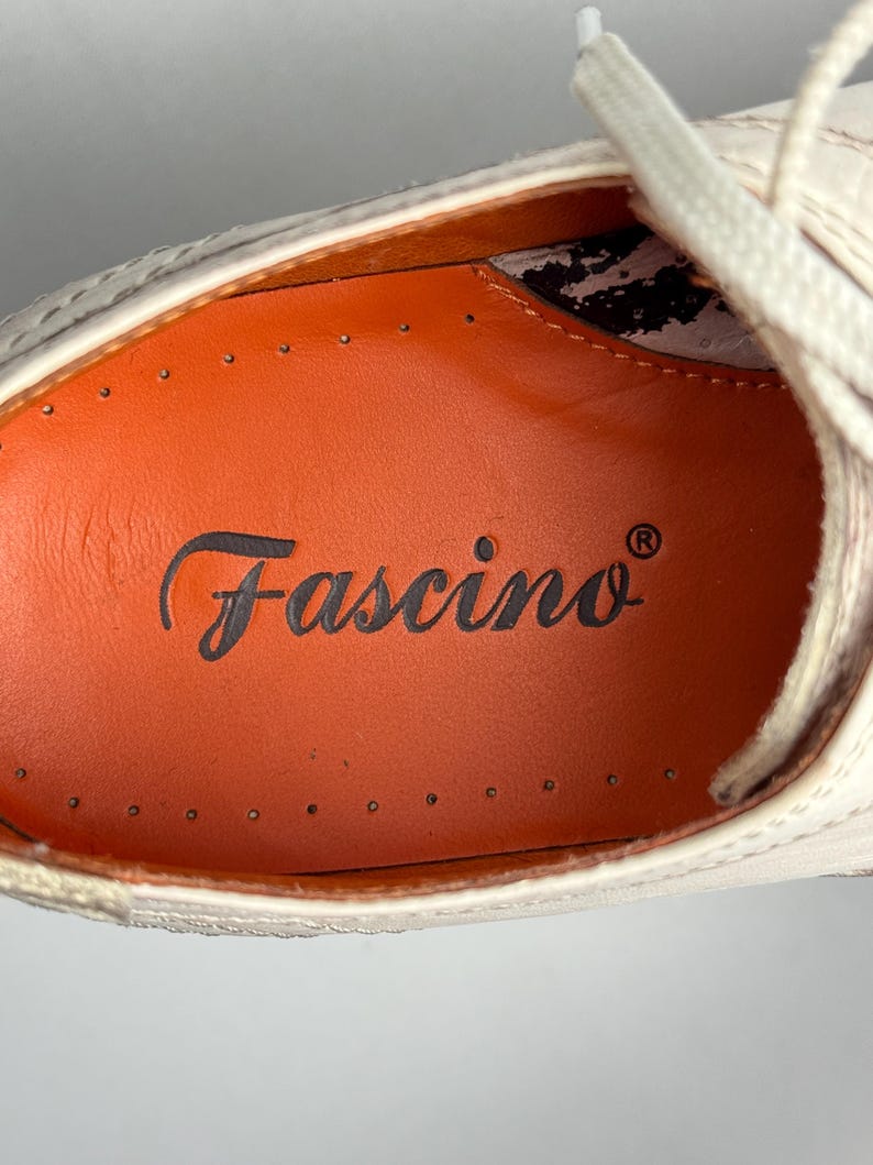 K&ouml;nnte beinhalten: Nahaufnahme eines cremefarbenen Schuhs mit wei&szlig;en Schn&uuml;rsenkeln. Der Schuh hat eine orangefarbene Innensohle mit dem Wort "Fascino" in Schreibschrift. Der Schuh scheint aus Leder zu sein und hat dekorative N&auml;hte.