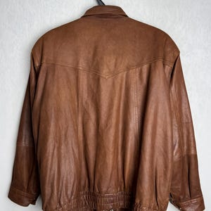 May include: A brown leather jacket with a classic design, hanging on a black hanger. The jacket features a collared neckline, a zipper closure, and elasticized cuffs and hem. The leather has a rich, textured appearance.