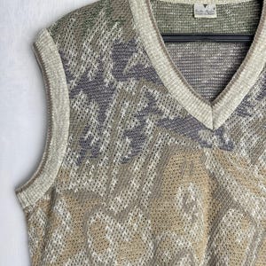 May include: A sleeveless sweater vest with a V-neck design. The vest features a patterned design in shades of beige, gray, green, and burgundy. The edges are trimmed with a light beige color. The vest is hanging on a black hanger.