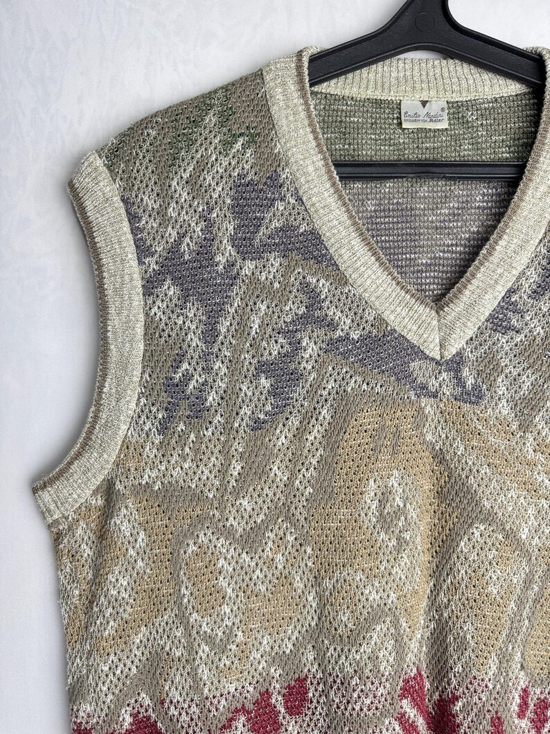 May include: A sleeveless sweater vest with a V-neck design. The vest features a patterned design in shades of beige, gray, green, and burgundy. The edges are trimmed with a light beige color. The vest is hanging on a black hanger.