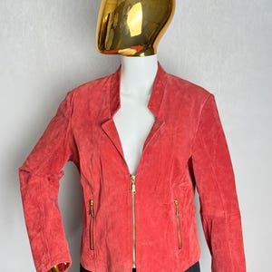 Puede incluir: Chaqueta de ante color coral con cuello alzado y cremallera completa. La chaqueta tiene dos bolsillos con cremallera y herrajes dorados. La chaqueta se exhibe en un maniqu&iacute;.