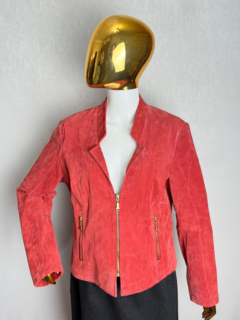 Puede incluir: Chaqueta de ante color coral con cuello alzado y cremallera completa. La chaqueta tiene dos bolsillos con cremallera y herrajes dorados. La chaqueta se exhibe en un maniqu&iacute;.