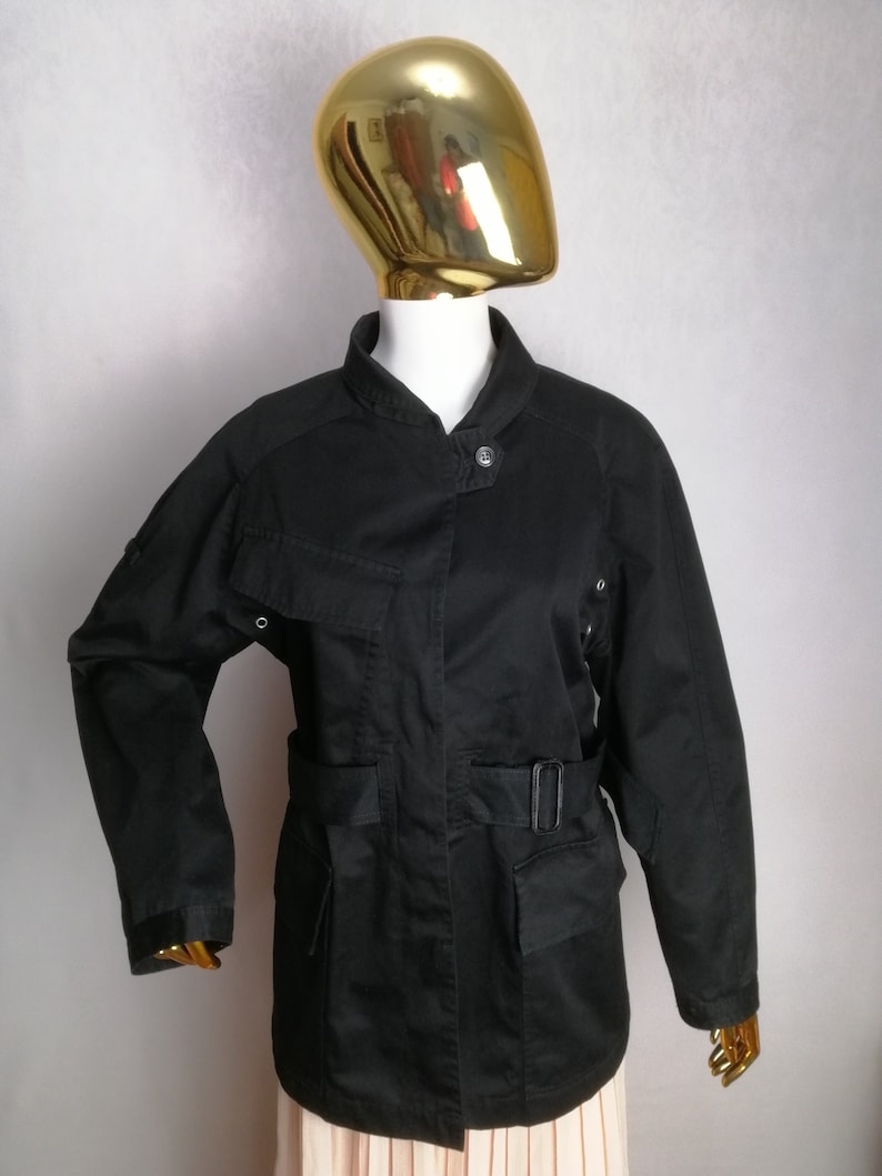 May include: A black utility jacket with a belted waist, multiple pockets, and a stand-up collar.