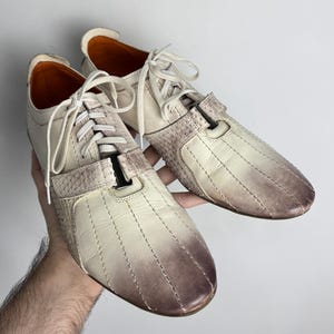 K&ouml;nnte beinhalten: Ein Paar cremefarbene Ledersneaker mit braunem Farbverlauf an Zehen und Ferse. Die Schuhe haben wei&szlig;e Schn&uuml;rsenkel und einen strukturierten Einsatz an der Seite. Die Innenseite ist orange.