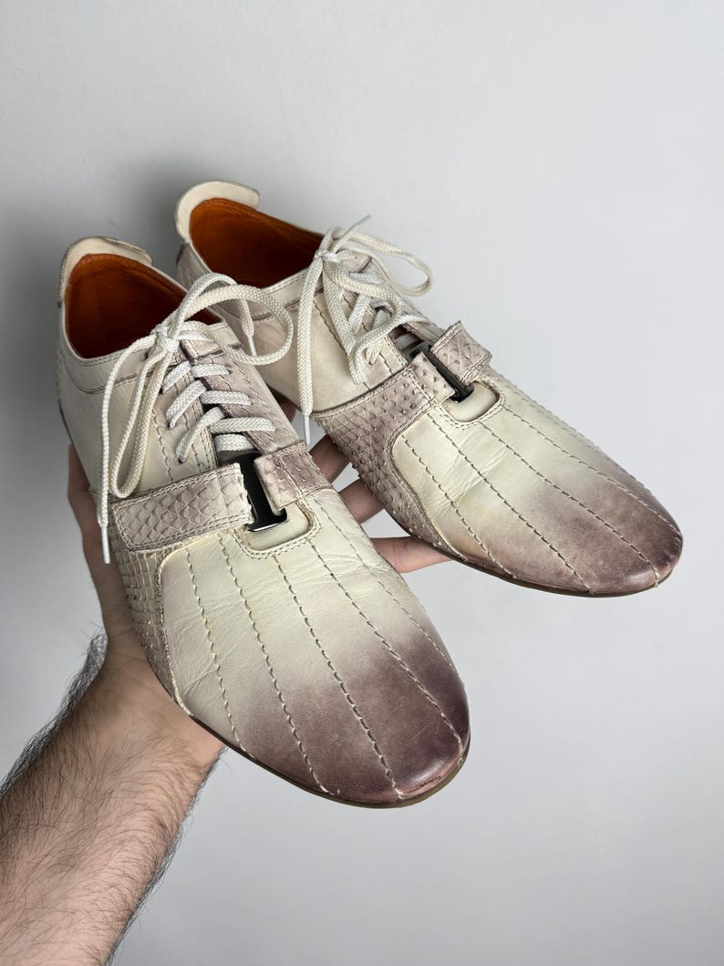 K&ouml;nnte beinhalten: Ein Paar cremefarbene Ledersneaker mit braunem Farbverlauf an Zehen und Ferse. Die Schuhe haben wei&szlig;e Schn&uuml;rsenkel und einen strukturierten Einsatz an der Seite. Die Innenseite ist orange.
