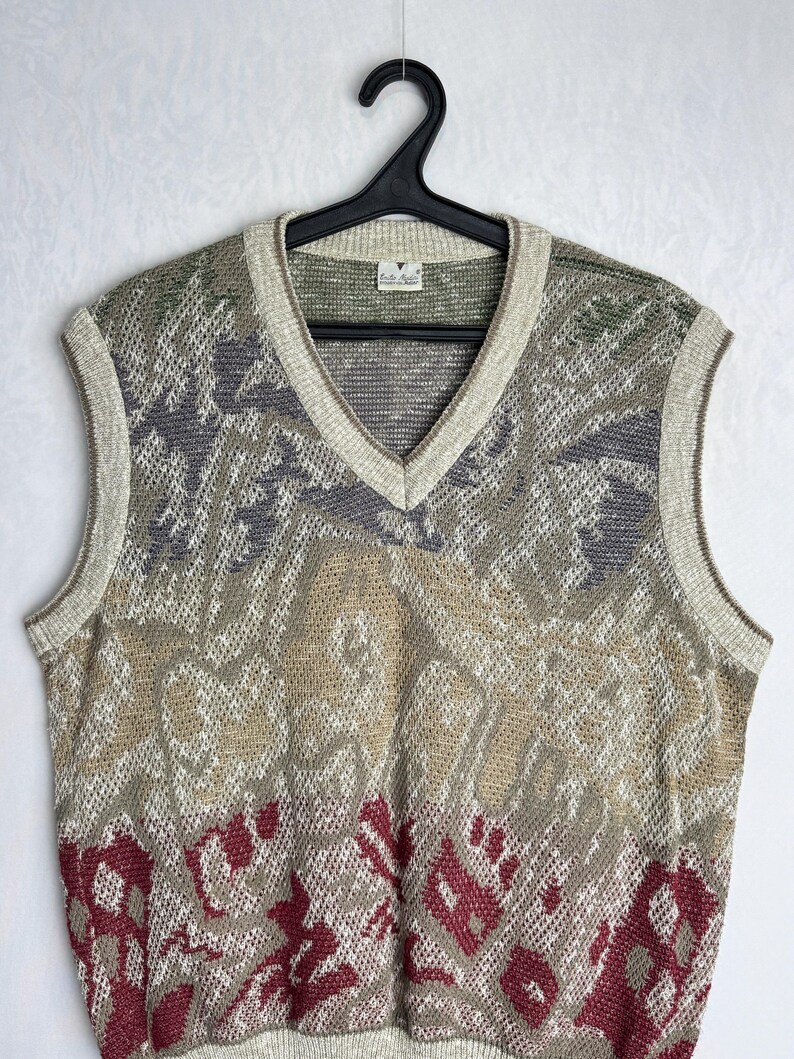 May include: A vintage V-neck sweater vest with a patterned design in shades of green, gray, beige, and burgundy. The vest is sleeveless and has a ribbed trim around the neckline and armholes. The garment is hanging on a black hanger.