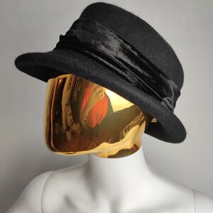May include: A black hat with a rounded crown and a small brim. A wide, black velvet band wraps around the base of the crown. The hat is displayed on a gold mannequin head against a gray background.