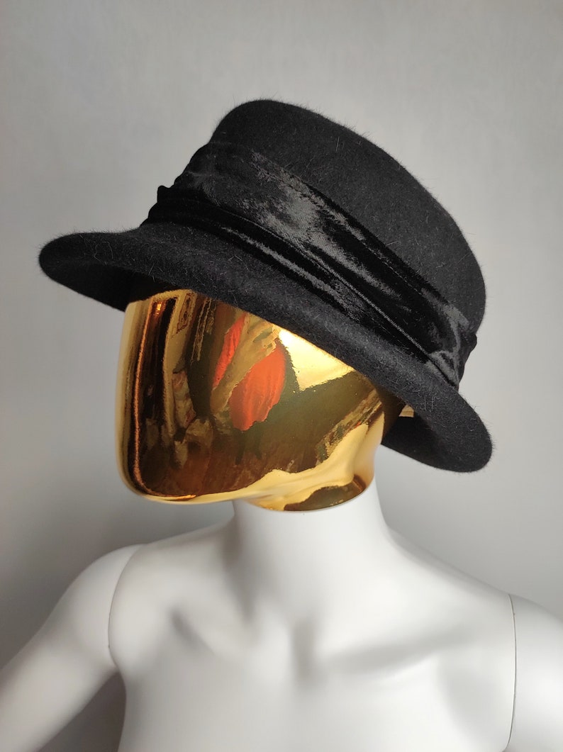 May include: A black hat with a rounded crown and a small brim. A wide, black velvet band wraps around the base of the crown. The hat is displayed on a gold mannequin head against a gray background.