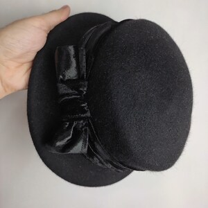 May include: A black hat with a rounded crown and a wide brim. The hat features a velvet bow on the side. The hat is made of a felt-like material and is suitable for formal occasions.