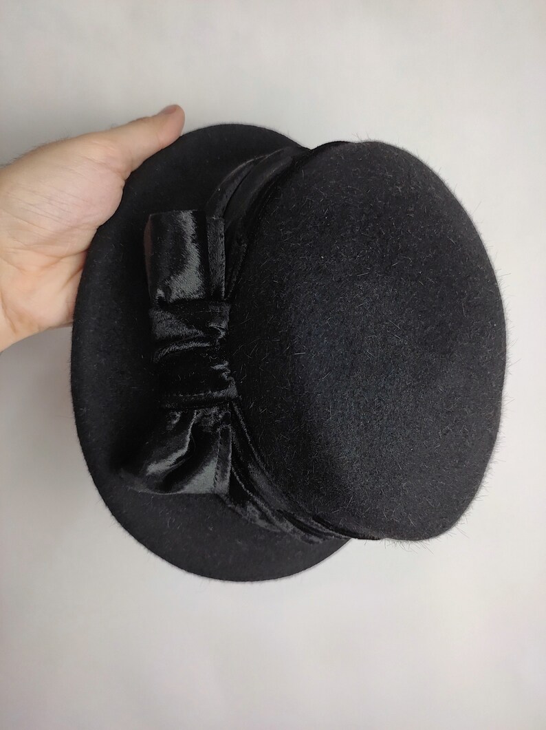 May include: A black hat with a rounded crown and a wide brim. The hat features a velvet bow on the side. The hat is made of a felt-like material and is suitable for formal occasions.