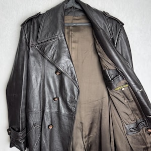 May include: A dark brown leather trench coat with a double-breasted design, hanging on a black hanger. The coat features a notched collar, shoulder epaulets, and buttoned cuffs. The interior lining is a contrasting brown color.