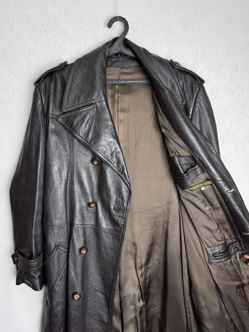 May include: A dark brown leather trench coat with a double-breasted design, hanging on a black hanger. The coat features a notched collar, shoulder epaulets, and buttoned cuffs. The interior lining is a contrasting brown color.