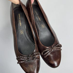 Puede incluir: Un par de bailarinas de cuero marr&oacute;n con un detalle de lazo adornado con peque&ntilde;os tachuelas plateadas. Los zapatos tienen una punta redondeada y un tac&oacute;n bajo. La marca "Pierre Cardin" es visible en la plantilla.