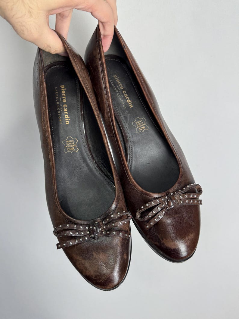 Puede incluir: Un par de bailarinas de cuero marr&oacute;n con un detalle de lazo adornado con peque&ntilde;os tachuelas plateadas. Los zapatos tienen una punta redondeada y un tac&oacute;n bajo. La marca "Pierre Cardin" es visible en la plantilla.