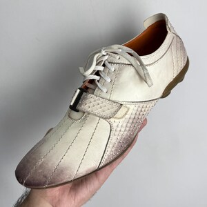 K&ouml;nnte beinhalten: Cremefarbener Ledersneaker mit strukturiertem, schuppenartigem Muster an Zehen und Ferse. Der Schuh hat wei&szlig;e Schn&uuml;rsenkel, einen Klettverschluss und eine braune Sohle. Das Leder hat einen Farbverlauf.