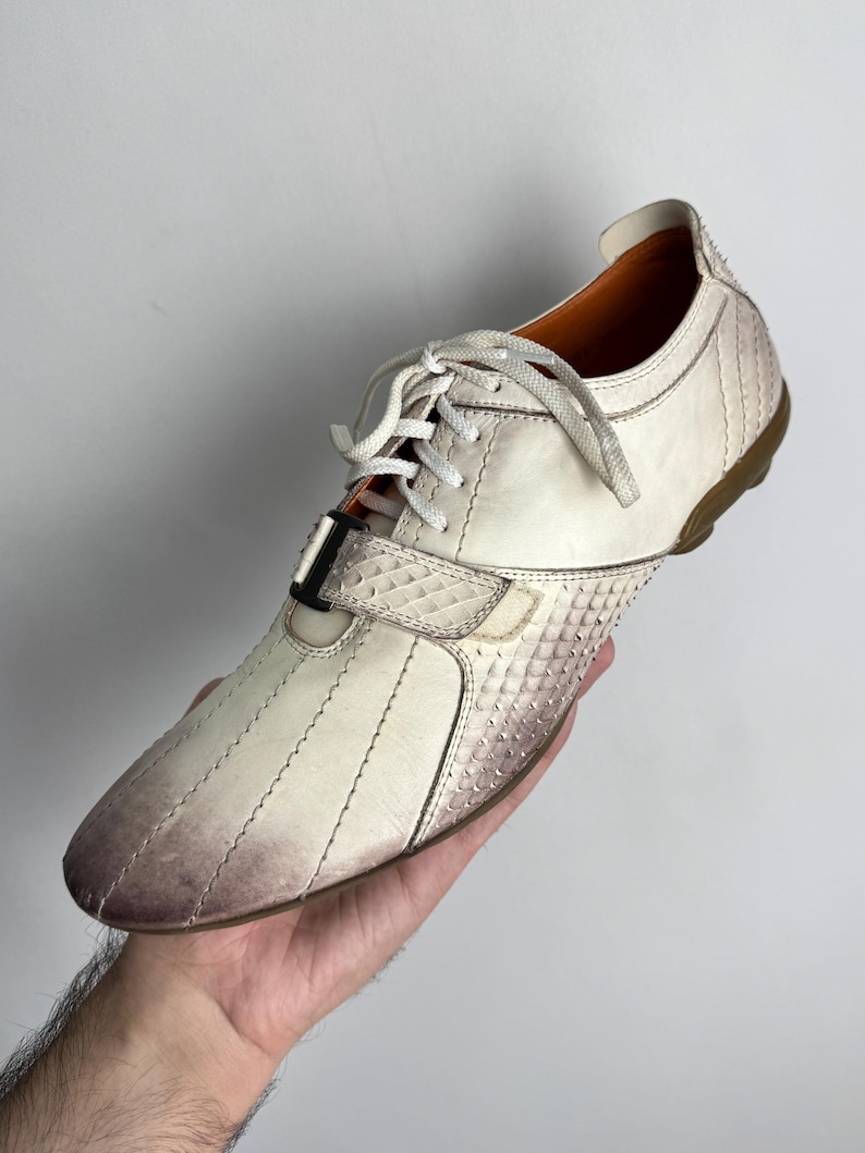K&ouml;nnte beinhalten: Cremefarbener Ledersneaker mit strukturiertem, schuppenartigem Muster an Zehen und Ferse. Der Schuh hat wei&szlig;e Schn&uuml;rsenkel, einen Klettverschluss und eine braune Sohle. Das Leder hat einen Farbverlauf.